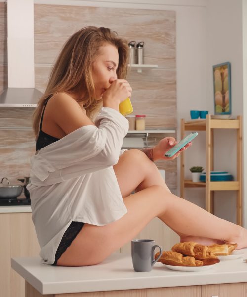 Seductive blonde housewife texting and sipping hot coffee wearing black lingerie sitting on kitchen table in the morning. Provocative young woman with tattoos in seductive underwear relaxing.